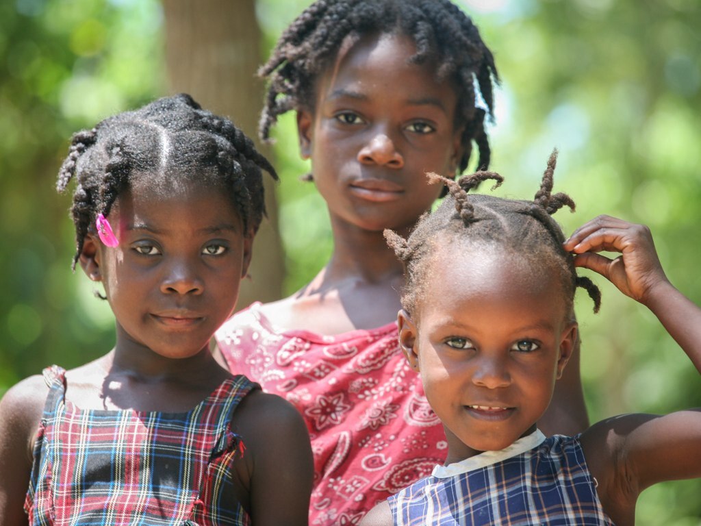 Three young Haitian girls are standing together outdoors with blurred greenery in the background. The girl on the left has short, curly hair with a pink clip and is wearing a plaid dress. The girl in the middle, who is the tallest, has longer, curly hair and is wearing a red patterned dress. The girl on the right has her hair styled in small braids and is wearing a blue plaid dress; she is smiling and touching her hair with one hand. They all have a calm and serene expression, and the natural light highlights their faces.