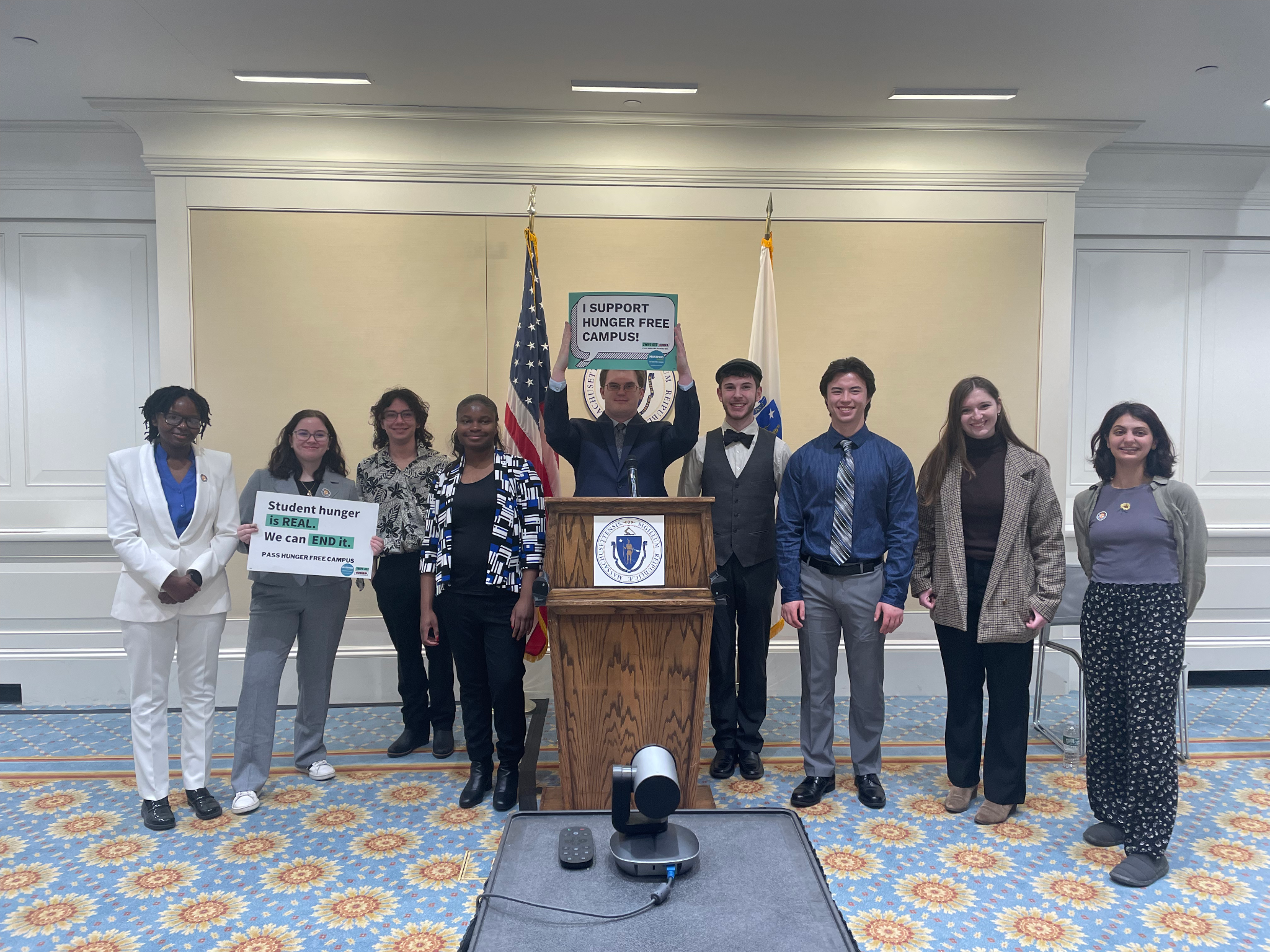 A group of nine people stands in a conference room with an American flag and the Massachusetts state flag behind them. They are posing for a photo, and most are smiling. The group includes individuals of different ages and ethnic backgrounds. Two people in the front row are holding signs that read, "Student hunger is REAL. We can END it. PASS HUNGER FREE CAMPUS" and "I SUPPORT HUNGER FREE CAMPUSES." The setting appears to be an official or formal environment, possibly a government building, indicated by the podium with a state seal. The room is decorated with a blue and floral patterned carpet and white paneling on the walls.