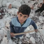 A young boy with dark hair and serious expression stands amidst a field of broken white rocks. He holds a hammer in his hand, wearing a blue sweater with a diamond pattern. Another person, partially visible, is also holding a hammer in the background. The image conveys a sense of labor and hardship.