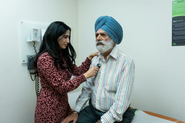 A young female doctor in a patterned dress is using a stethoscope to examine an elderly man wearing a blue turban and a striped shirt. They are in a medical examination room with medical equipment and a sign on the wall in the background.