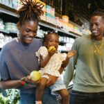 "An African American family is grocery shopping. The mother, holding their young daughter, is smiling while the child bites into a green apple. The father, standing next to them, is also smiling. They are in the produce section with shelves of vegetables and packaged goods in the background."