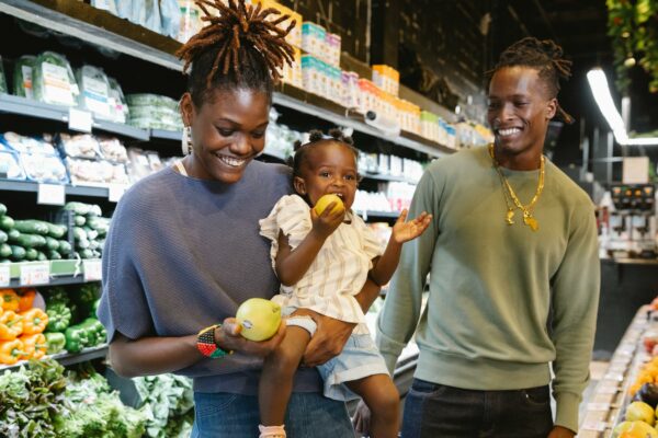 "An African American family is grocery shopping. The mother, holding their young daughter, is smiling while the child bites into a green apple. The father, standing next to them, is also smiling. They are in the produce section with shelves of vegetables and packaged goods in the background."