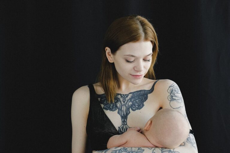 A young mother with a large tattoo of a ram's skull on her chest and floral tattoos on her arm is holding and breastfeeding her baby. She has light brown hair and is looking down lovingly at the baby. The background is black.