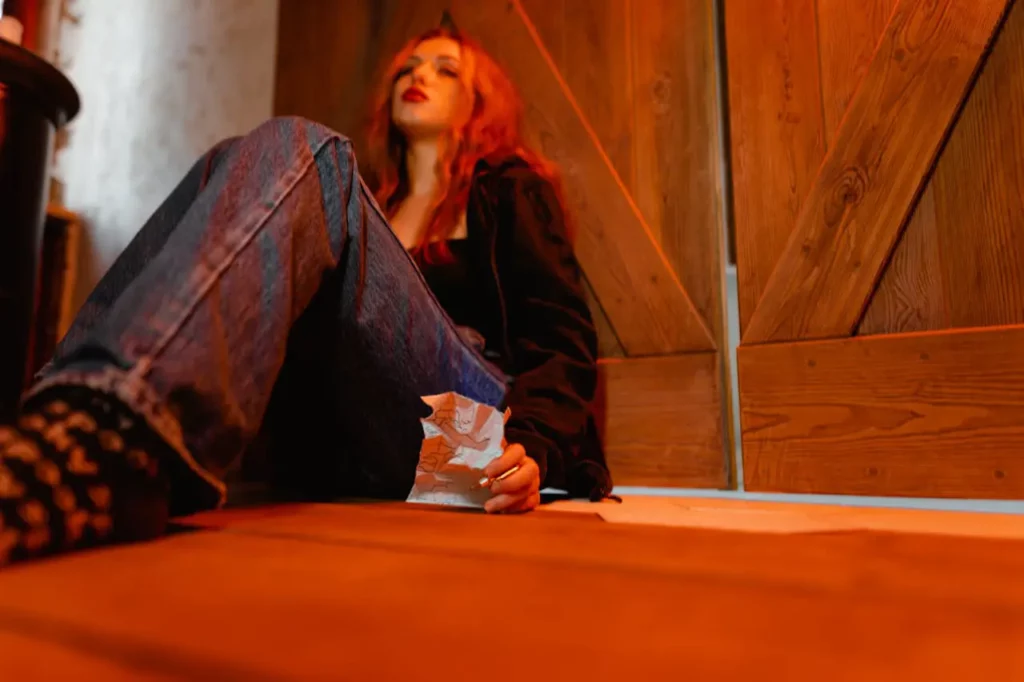 A young person with long red hair sits on the floor against a wooden wall, looking contemplative. They are wearing a dark jacket and jeans, holding a crumpled piece of paper in one hand. The warm, orange lighting creates a dramatic and introspective atmosphere, highlighting the mood of the scene.