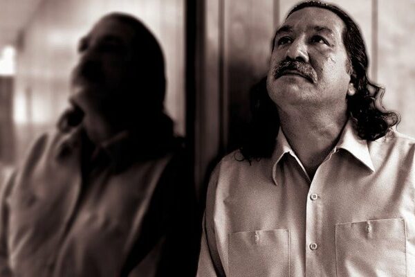 Photo of a Native American man, Leonard Peltier, standing with his side against a mirror, gazing upwards.