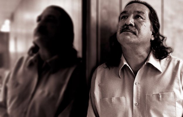 Photo of a Native American man, Leonard Peltier, standing with his side against a mirror, gazing upwards.